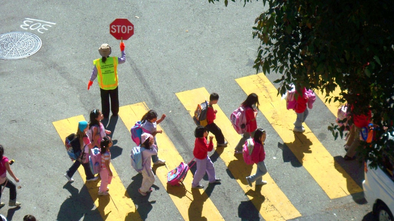 Kids crossing the street in a yellow crosswalk with a crossing guard, seen from above.
