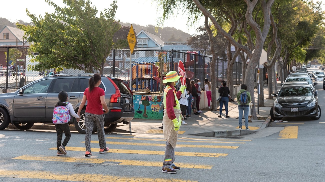 Crossing guard holding up a stop sign as parents and kids walk to school in the background.