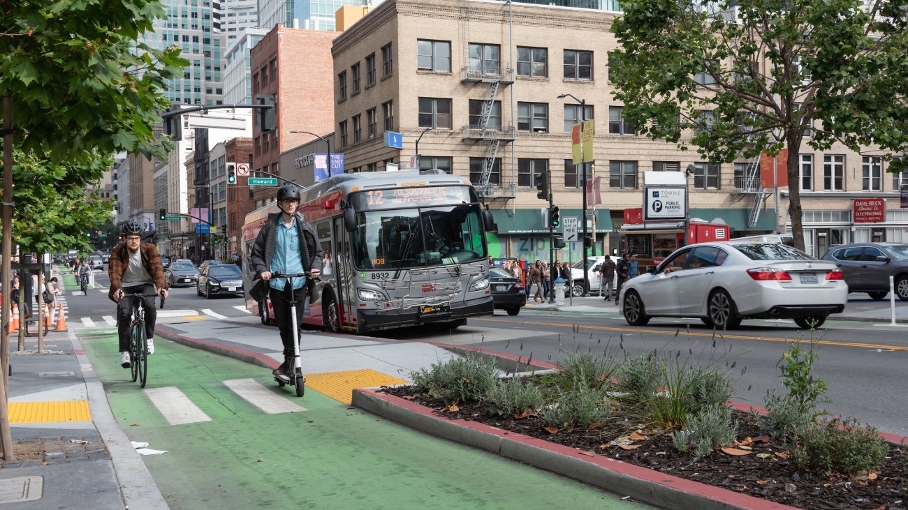 people on bike and scooter on green lane, transit boarding island with landscaping, and Muni bus