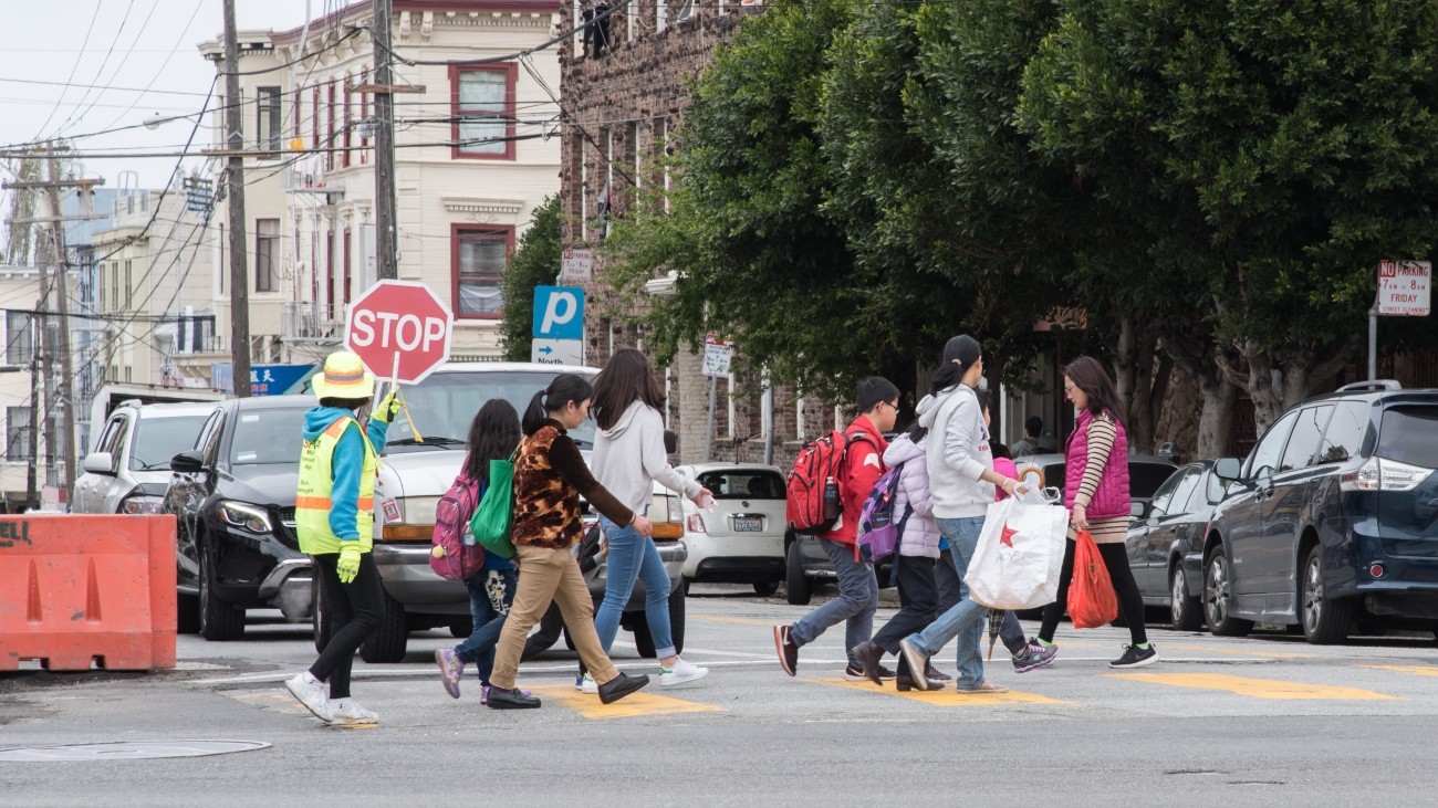 Children and adults crossing the street with a crossing guard along Broadway Street