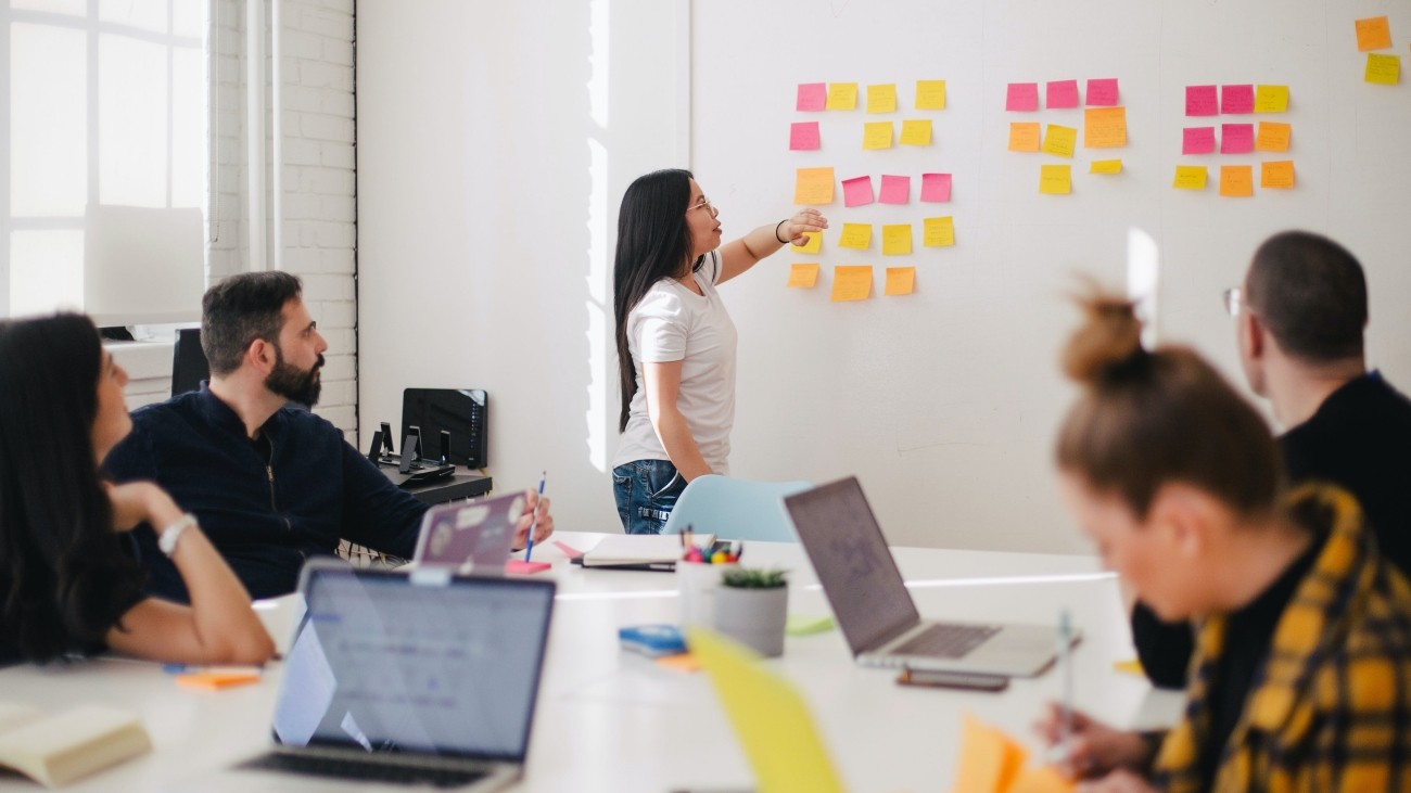 People sitting in front of laptops around a table, person standing at wall and pointing to post it notes