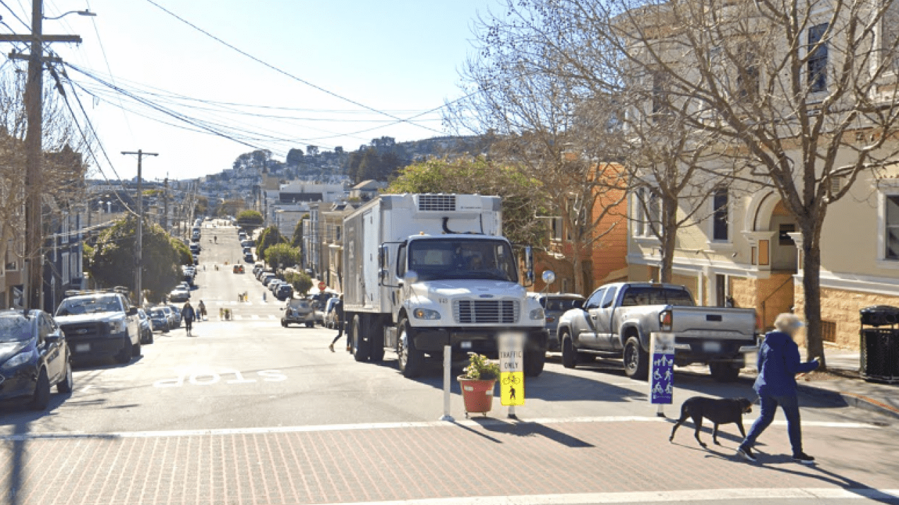 Street-level view of Sanchez Slow Street on a sunny day. A person walks a dog across the intersection.