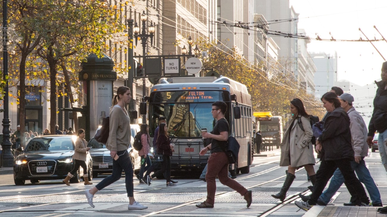 Pedestrians crossing the street with a Muni bus and vehicles waiting at the intersectionn