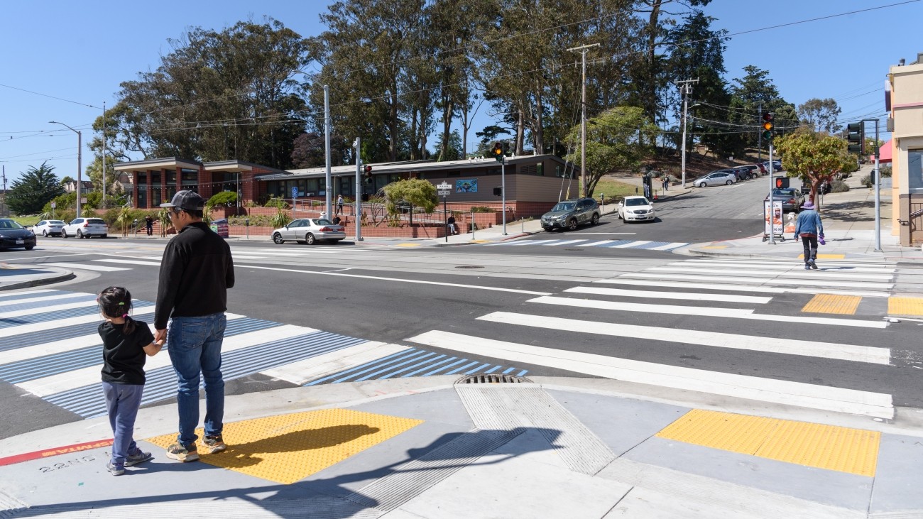 An adult and a child holding hands waiting on the sidewalk with a curb ramp, by a decorated high visibility crosswalk across from the Parkside Library along Taraval Street