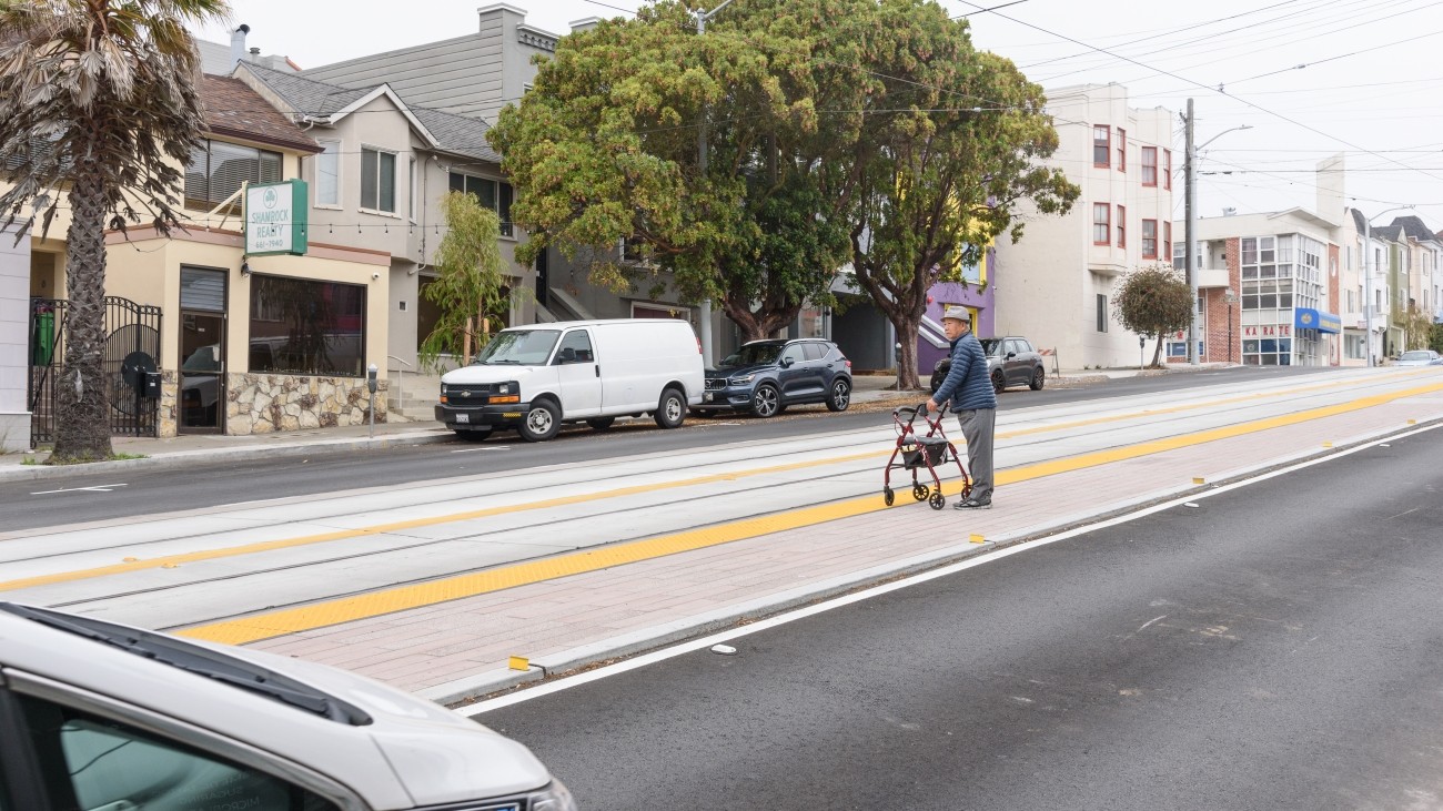 A senior citizen wearing a hat holding a walker waiting for L Taraval at new boarding  platform on Taraval Street