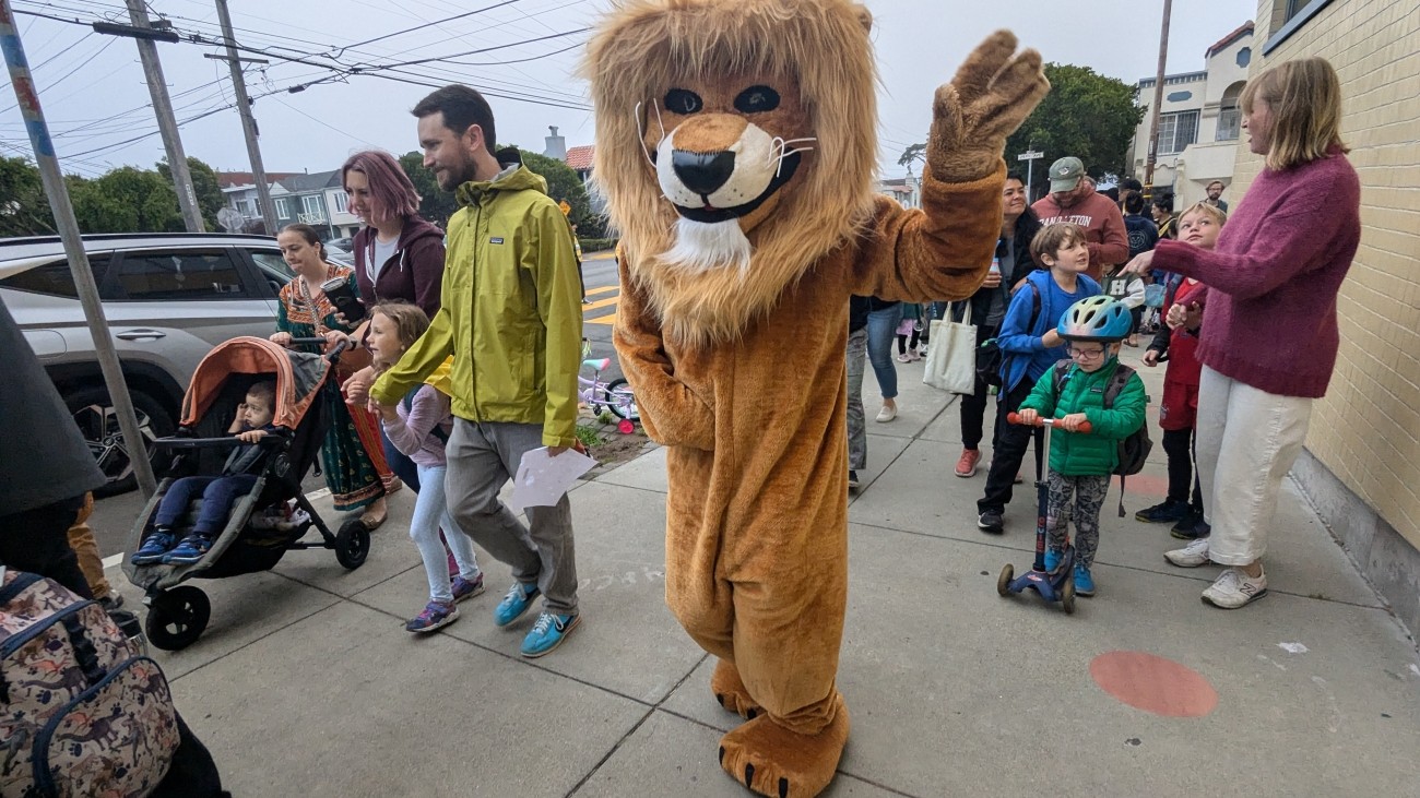 Dianne Feinstein Elementary's school mascot waving and celebrating walk and roll to school day, with parents and students in the background