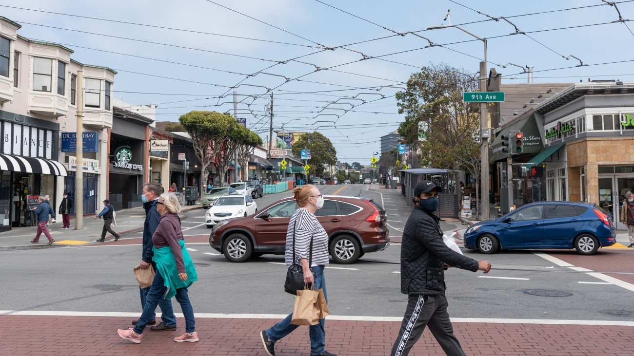 Pedestrians crossing the street at 9th Avenue and Irving Street, vehicles and businesses in the background