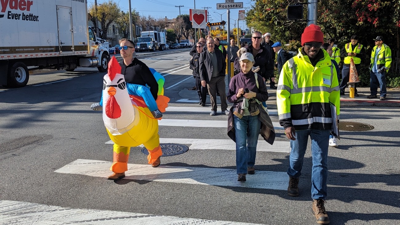 People walking in a crosswalk, with one person wearing a costume of a chicken