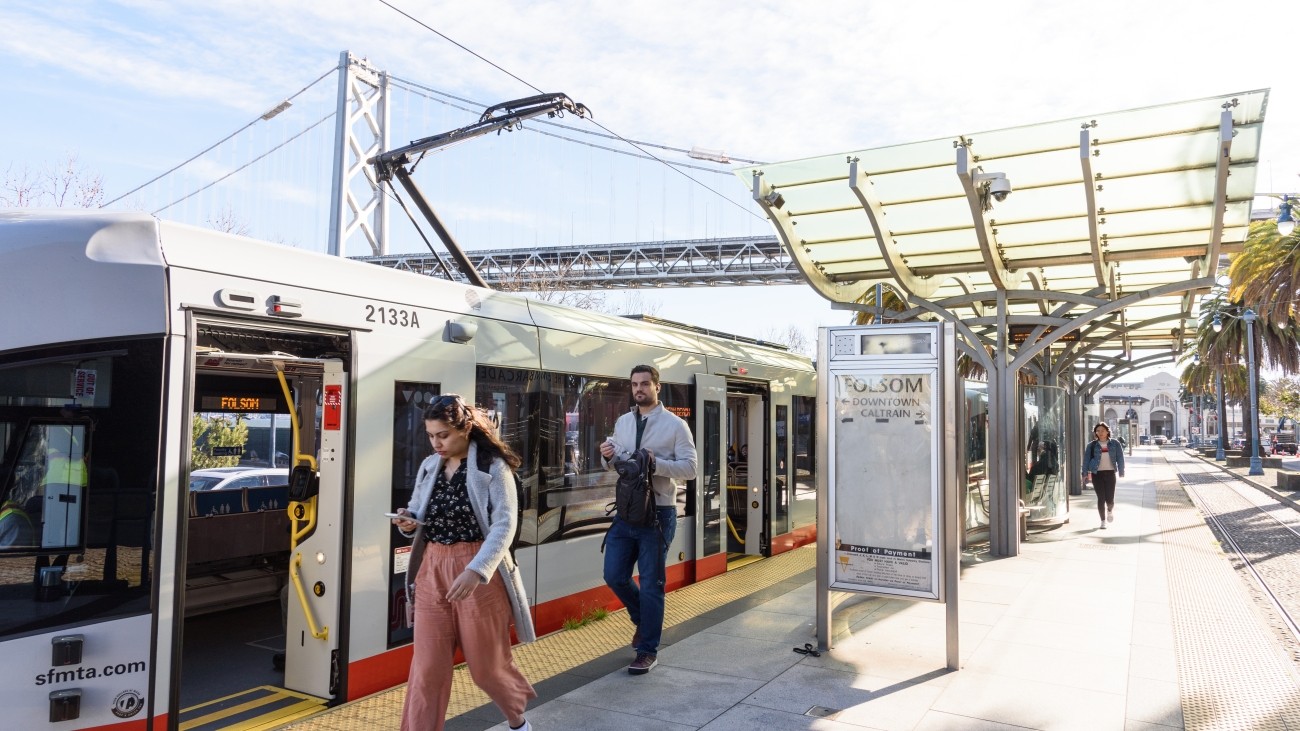 People offboarding a Muni metro train onto a boarding platform, with the Bay Bridge in the background