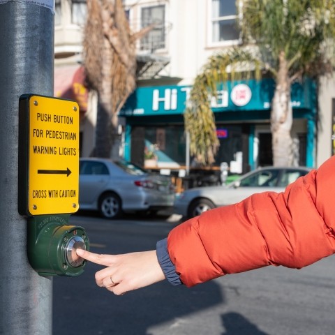 A person with an orange jacket pressing a button to activate pedestrian warning lights at a crosswalk