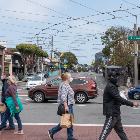 Pedestrians crossing the street at 9th Avenue and Irving Street, vehicles and businesses in the background