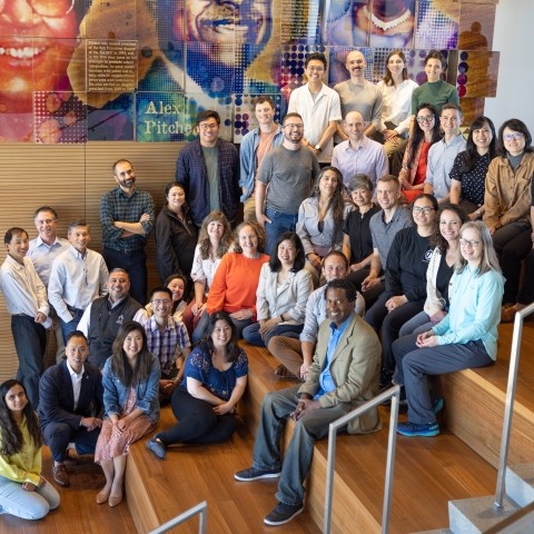 A few dozen individuals seated on a staircase smiling looking at the camera. There is a mural type design on the backwall.   